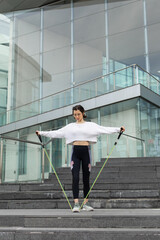 Young sporty woman doing lateral raises for exercising shoulders with resistance band. Urban fitness outdoor workout.