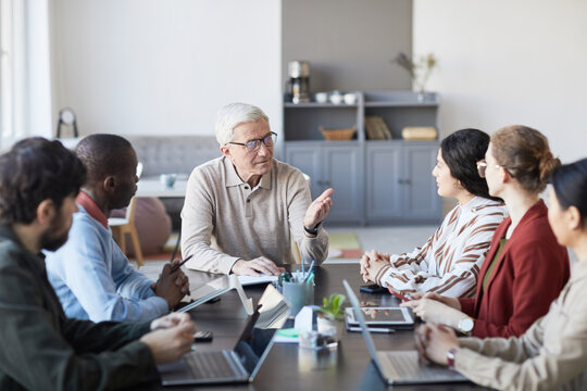 Portrait Of Modern Senior Businessman Talking To Group Of People During Meeting At Table In Office