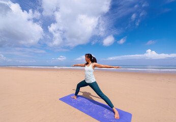 people, fitness, sport and healthy lifestyle concept - asian woman making yoga warrior pose on tropical beach and blue sky background