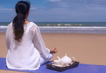 people and leisure concept- young asian woman meditating and drinking tea on the tropical beach