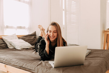 Smiling young business woman freelance student in domestic pajamas working remotely using technology and talking on a mobile phone looking at a laptop sitting in bed at home, selective focus