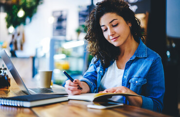 Caucaisan female student writing education notes in textbook while e learning in cafe interior,...