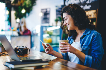 Happy hipster girl with coffee to go using cellular device for online messaging and chatting in...