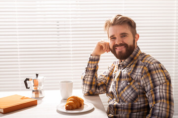 Pensive young bearded male businessman drinking cup of coffee on background of blinds. Concept of pleasant morning or lunch break.