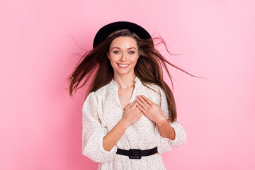 Photo of shiny thankful young woman dressed white clothes cap arms chest air blowing isolated pink color background