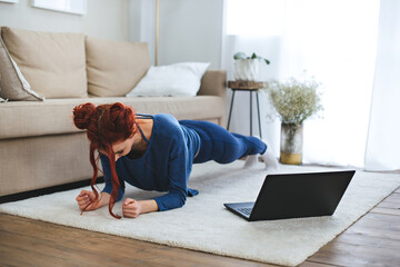 A young woman sits on a carpet near a laptop doing yoga at home. Staying home.