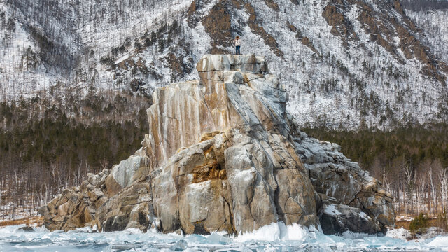 A large rock with steep slopes rises above the icy and hummocky lake. A man in a down jacket stands on a flat top. The background is snow-capped and wooded mountains. Lake Baikal. Russia