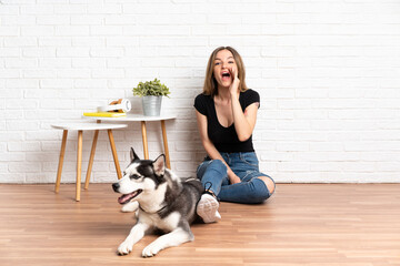 Young pretty woman with her husky dog sitting in the floor at indoors shouting with mouth wide open