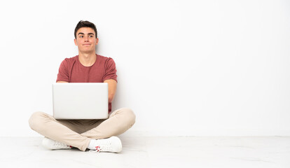 Teenager man sitting on the flor with his laptop making doubts gesture while lifting the shoulders