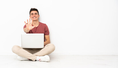 Teenager man sitting on the flor with his laptop counting five with fingers