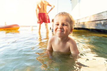 Cute adorable cheerful caucasian little blond toddler boy enjoy having fun playing at lake or river beach water on warm sunset evening time outdoors.. Happy childhood vacation at countryside concept