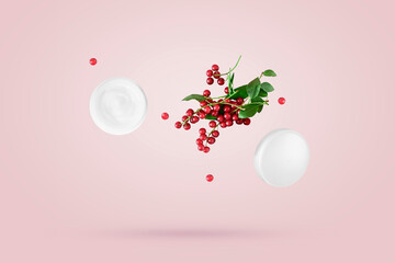 An open jar of cosmetic cream and a bunch of small red berries float freely in the air against a pink background. Levitation concept, natural ingredients for cosmetic products.