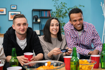 A multinational group of college students sit on the couch and play video games on the TV at home. Smiling friends hang out. A party of teenage students