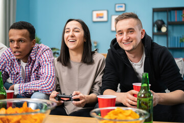 The smiling friends spend their free time playing video games on a console while sitting on the couch in the living room at home. A cheerful student girl wins a round
