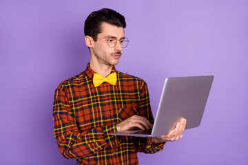 Photo of focused young guy hold laptop look screen wear eyewear bow tie checkered shirt isolated violet background