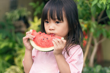 Portrait​ image​ of​ 2-3 yeas​ old​ of​ baby.​ Happy​ Asian child girl eating and biting a piece of watermelon. Enjoy eating moment. Healthy food and kid concept.​ Sucking fingers in the mouth.