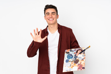 Teenager artist man holding a palette isolated on white background saluting with hand with happy...