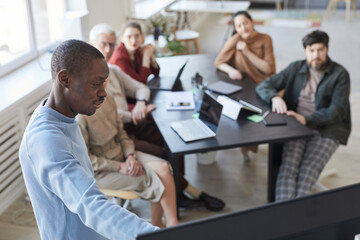 Obraz premium High angle portrait of African-American man giving presentation in office to diverse business team and pointing at digital screen, copy space