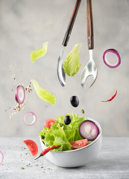 bowl with fresh salad, ingredients and cooking spoons in levitation on gray stone background