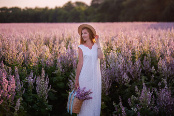 A young woman in a white sundress, straw hat holds a wicker basket with a bouquet. A girl walks through a sage blooming pink field at sunset. The concept of tenderness, beauty of untouched nature