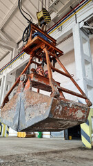 Rusty grab bucket or clamshell hanging on crane in empty industrial plant.
