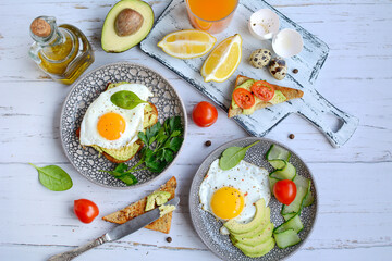 Two plates with fried eggs, toast, avocado, tomatoes, olive oil in a bottle, cutting board with lemon slices on a light background, top view.