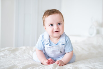 baby boy 6 months old in blu clothes smiling and sitting on white bed at home.