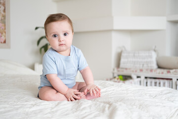 baby boy 6 months old in blu bodysuit smiling and sitting on white bed at home.