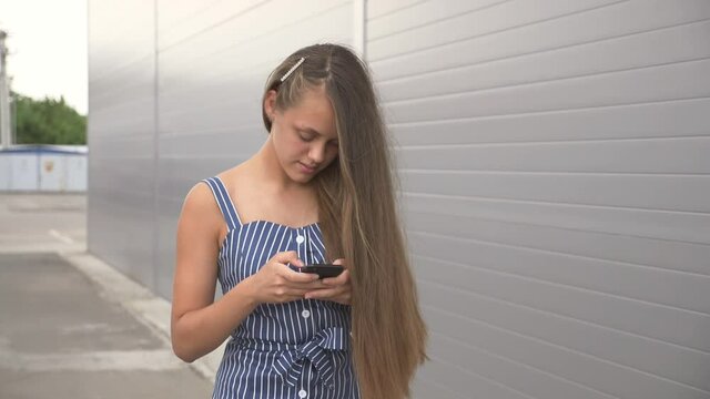 Smiling girl walking down street with mobile phone in her hands. girl looks into phone, types on keyboard text. teenager uses smartphone to communicate. teenager walking down street looks at the phone