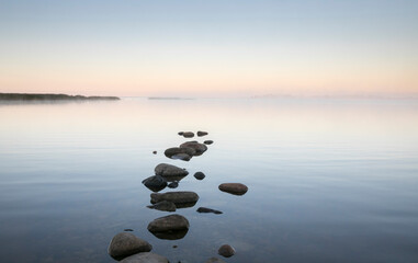 Beautiful sea bay scenery with the row of stones on the calm sunrise colored water and the fog...