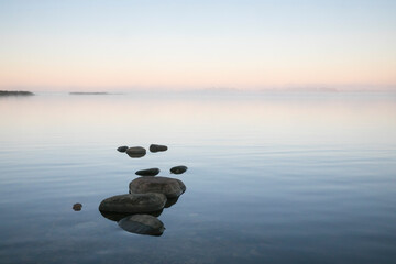 Beautiful sea bay scenery with the row of stones on the calm sunrise colored water and the fog partly hiding view to the sea islands in horizon