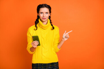 Photo of brown hair optimistic girl hold telephone point empty space wear yellow sweater isolated on orange color background