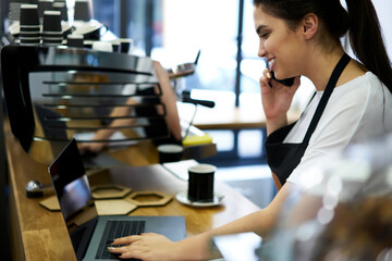 Positive female owner of small business cafe using devices for organizing job making call to banking service operator, smiling waitress talking on cellular with clients reserving table in coffee shop