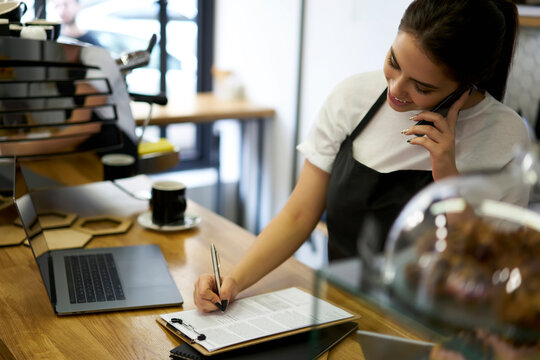 Smiling young woman owner of coffee shop making smartphone call filling information about franchise working with technology, prosperous female barista talking on mobile phone about bakery supply