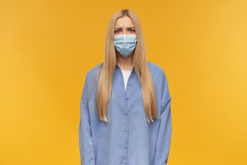 Frowning girl, unhappy looking woman with blond long hair. Wearing blue shirt and medical face mask. People and emotion concept. Watching at the camera, isolated over orange background