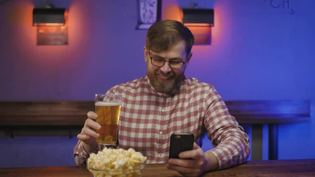 Portrait of laughing man with beard and in glasses seeing something funny on mobile phone in pub. happy millenial reading message at bar counter with beer and pocorn. laughing in hallway