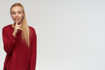 Teenage girl, happy looking woman with blond long hair. Wearing red sweater. People and emotion concept. Watching to the right at copy space, isolated over white background, show silence sign