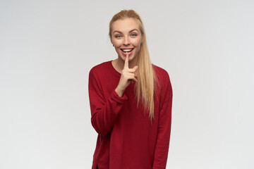 Teenage girl, happy looking woman with blond long hair. Wearing red sweater. People and emotion concept. Watching to the right at copy space, isolated over white background, show silence sign