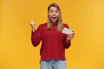 Pretty woman keeps mobile phone in her hand, clenched one fist looking with excitement, opened her mouth, like screaming wow, won the game or got a discount, isolated on a yellow studio background