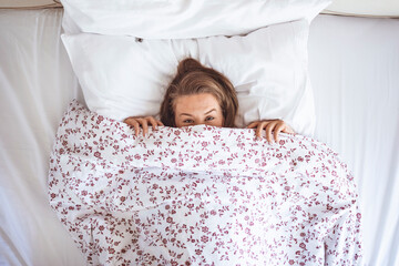 Funny young woman lying in bed and hiding under sheet while looking up with copy space. Top view of girl hiding face under white blanket on bed in the morning. 