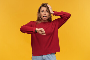 indoor shot of young woman standing over orange background, keeps her mouth open widely with a confused, scared facial expression, watching on her watches and keep her hand on her head.