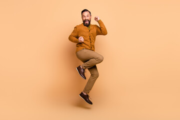 Fototapeta na wymiar Full size profile photo of optimistic brown hair man jump wear brown sweater pants shoes isolated on beige color background
