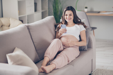 Full body photo of happy cute young woman hold smartphone read touch belly lie sofa inside house indoors