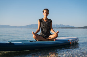 Fototapeta na wymiar Young woman meditating in lotus position sitting on a sup board