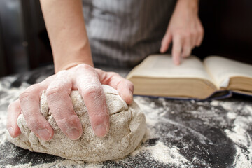 Baker holds one hand on the dough and the other on the cookbook, on the table sprinkled with flour....