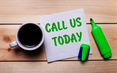 On a wooden table, a cup of coffee, a green marker and a napkin with the text CALL US TODAY