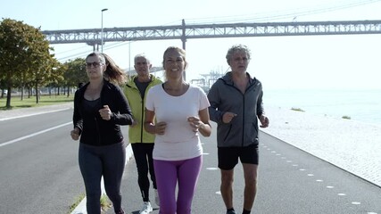 Active mature joggers running on promenade. Group of senior people in sporty clothes jogging outside together. Dolly shot. Elderly age and lifestyle concept - Powered by Adobe