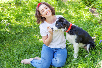Smiling young attractive woman playing with cute puppy dog border collie in summer garden or city park outdoor background. Girl training trick with dog friend. Pet care and animals concept.