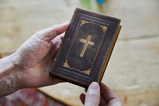 Old man's hands hold a Bible from the 19th century. A cross is printed on the holy scripture. Who will save you? The Bible gives security to many people. 