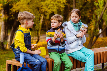 Two boys and girl have a snack on a bench in the park and chat. Autumn day.